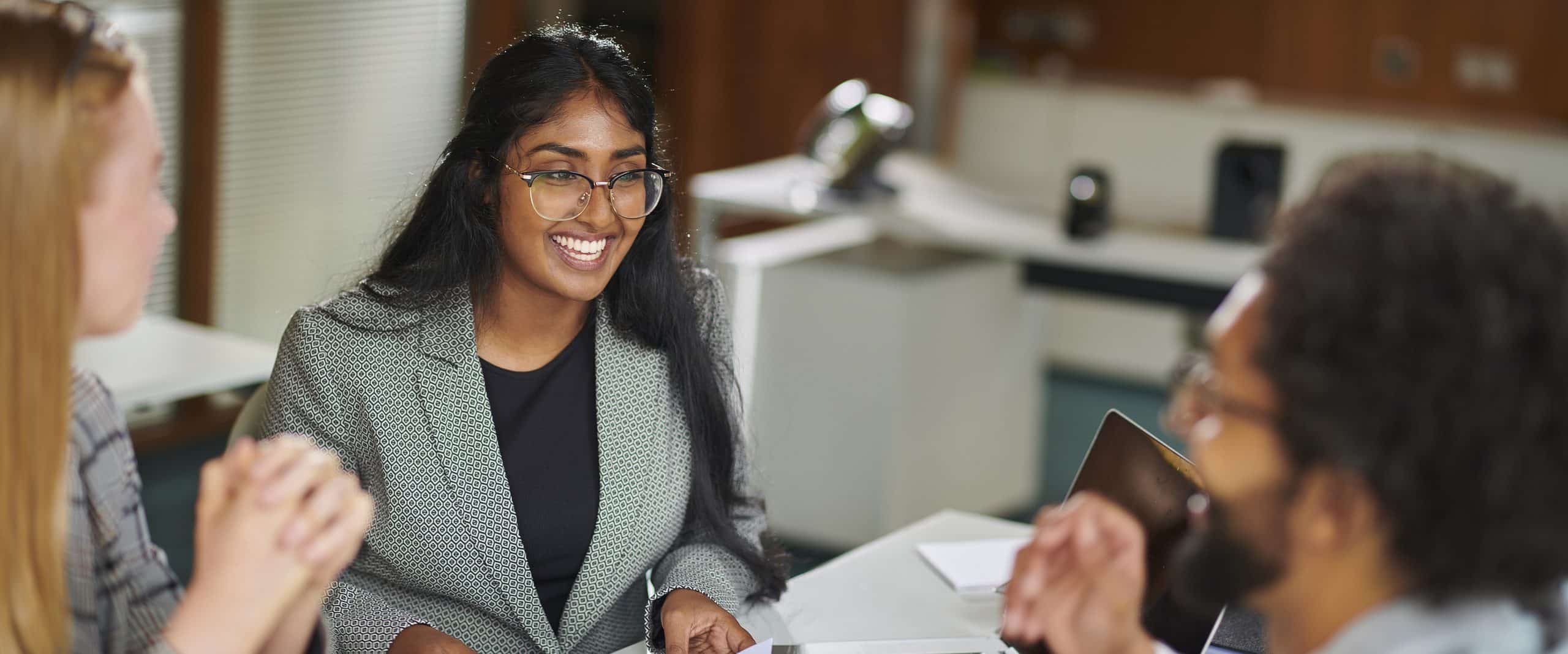 Three people sit around a table in a brightly lit office, engaged in conversation. One person in the center is smiling, holding a pen. Papers, a laptop, and office equipment are visible in the background.