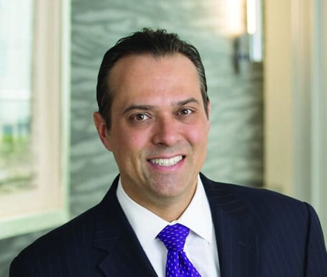 A man wearing a dark pinstripe suit, white shirt, and blue polka dot tie is smiling at the camera. He is indoors, standing in front of a blurred background with neutral tones and a wall sconce.