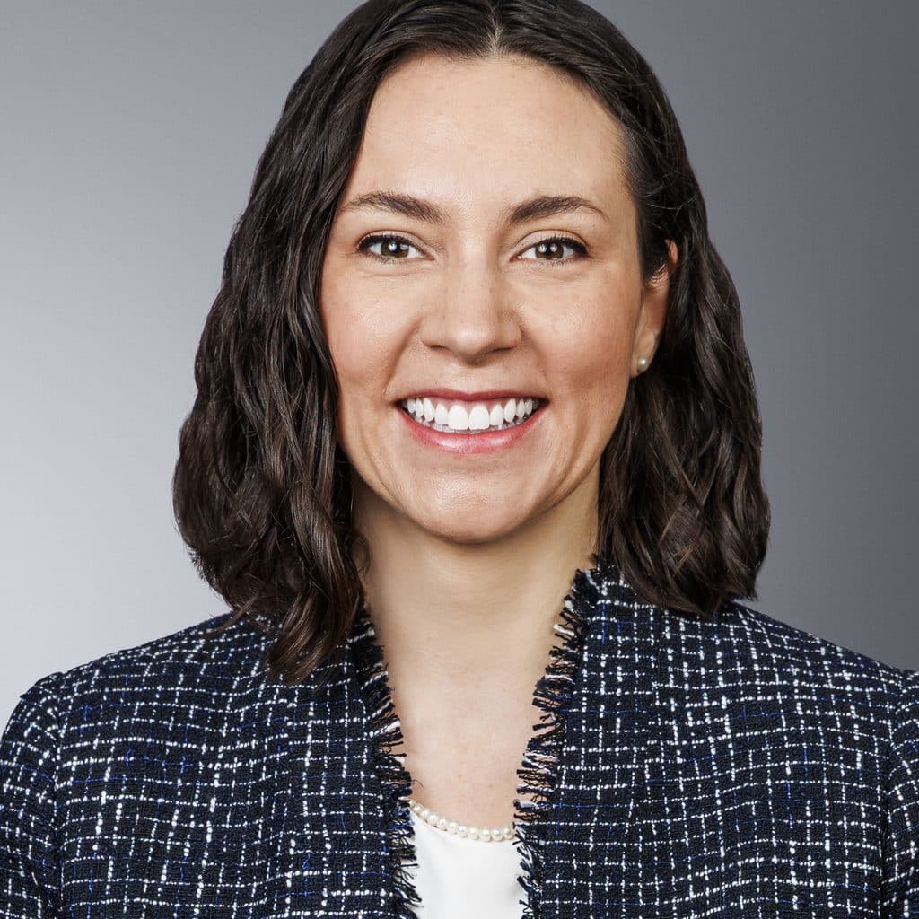 A woman with shoulder-length dark hair is smiling at the camera. She is wearing a navy textured blazer over a white top, with a string of pearls visible. The background is a neutral gray gradient.