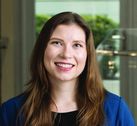 A woman with long brown hair wearing a blue blazer over a black top is smiling at the camera. There is a blurred indoor background with a window and greenery outside.