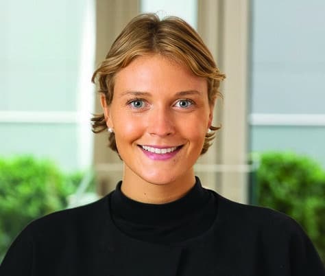 A person with short blonde hair, wearing a black top with large decorative buttons, stands indoors in front of windows with greenery visible outside. The individual is smiling at the camera.