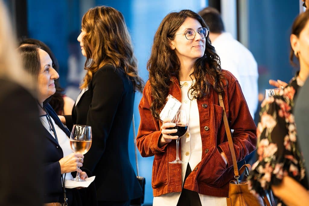 A woman with glasses and a corduroy jacket holds a glass of red wine while attending a social gathering.