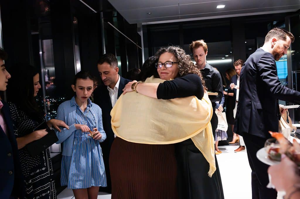 A woman wearing glasses and a yellow shawl hugs another person at an indoor gathering while others stand nearby.