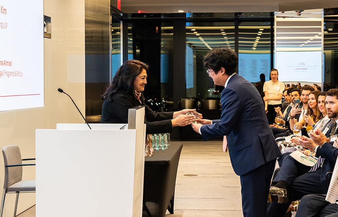 A woman presents an award to a man in a suit during a formal ceremony as an audience applauds.
