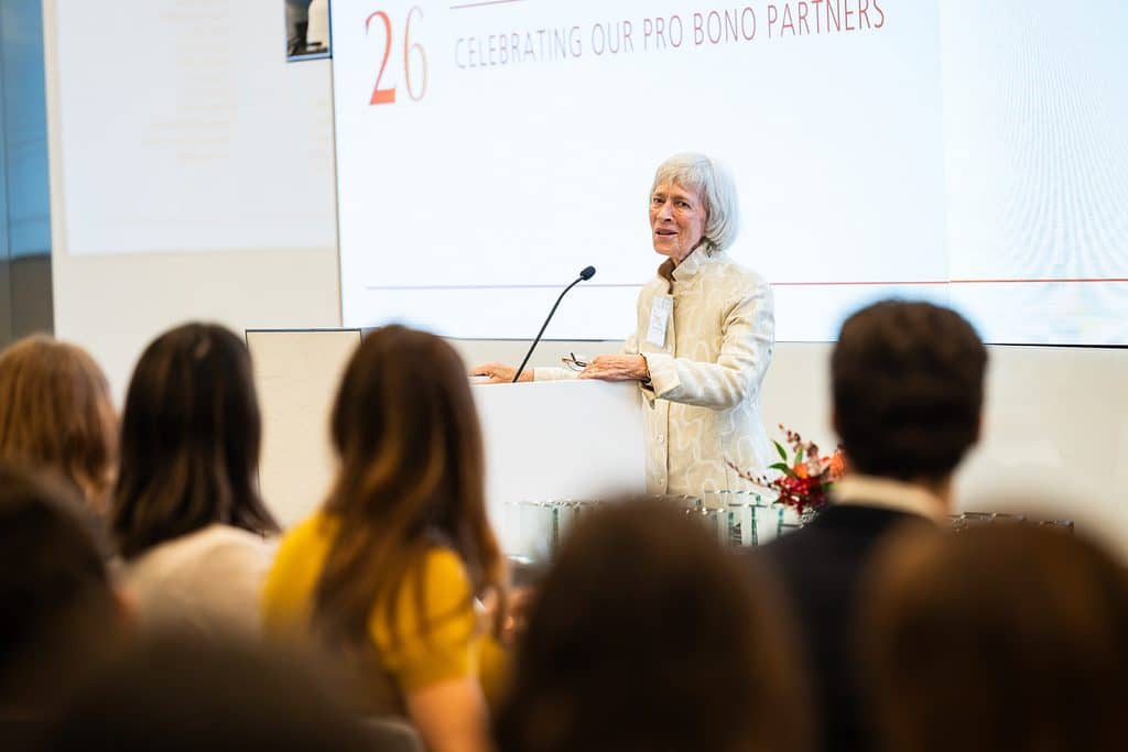 A woman stands at a podium speaking to an audience in front of a screen that reads 26 Celebrating Our Pro Bono Partners.