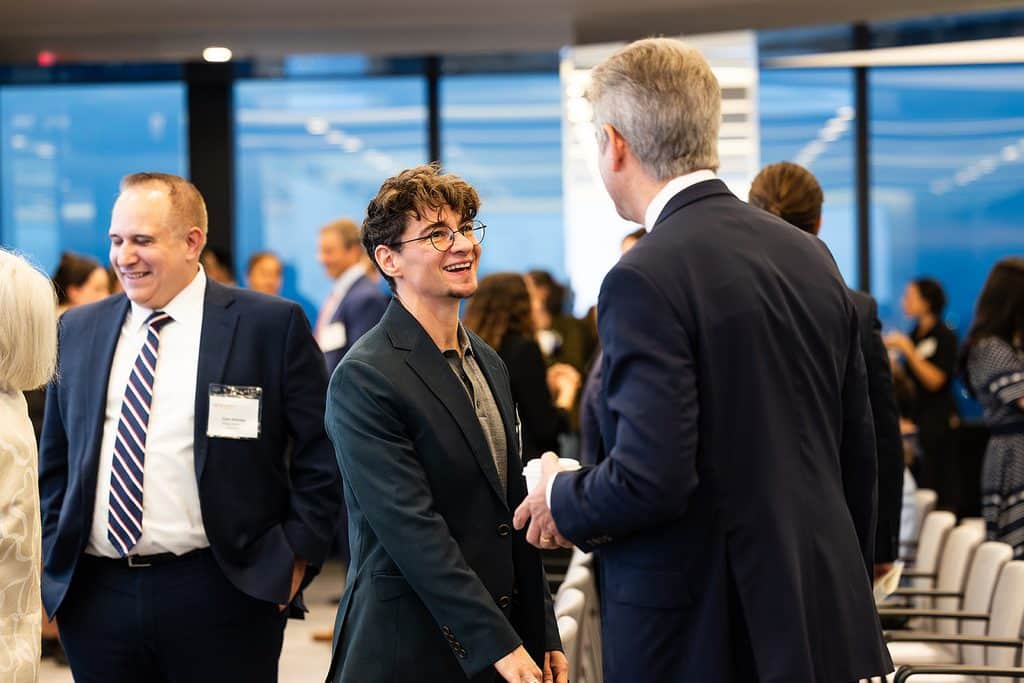 A young person with glasses smiles while talking to an older person in a suit at a professional networking event.