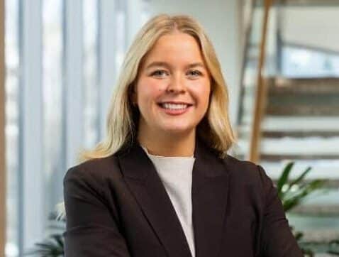A smiling blonde woman in a black blazer stands with her arms crossed in an office lobby.