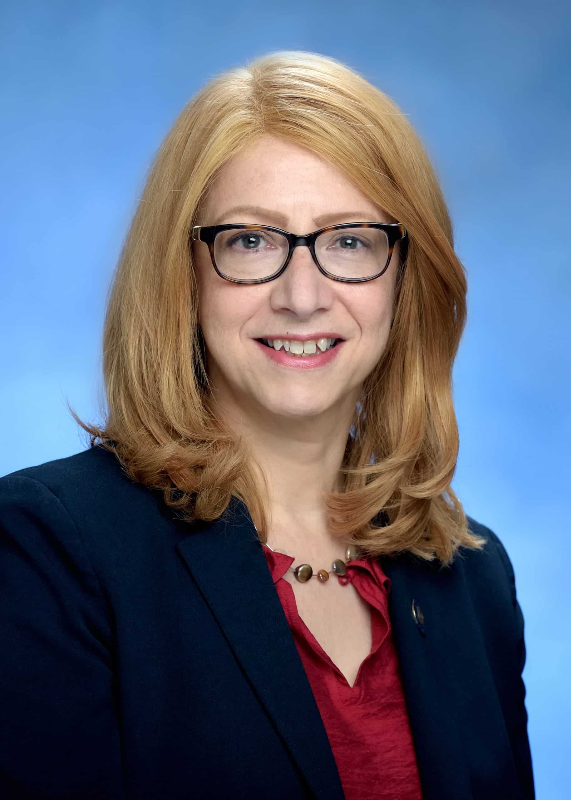 A smiling woman with shoulder-length blonde hair and glasses, wearing a navy blazer over a red blouse.